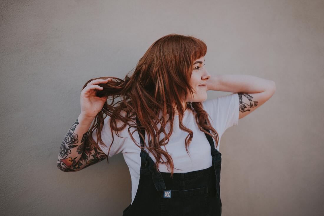 Portrait of a young woman with red hair and tattoos looking off camera and smiling. This represents that Sacred Circle Holistic Healing offers therapy in Kansas for Highly Sensitive People who are often women who view the world alternatively.