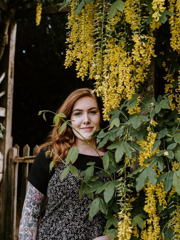 Woman with red hair and a tattoo standing next to yellow flowers. This illustrates that Sacred Circle Holistic Healing offers online counseling in Kansas for young adults.