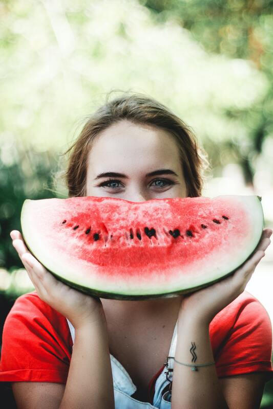 Young women holding a slice of watermelon in front of her smiling mouth. This illustrates how self-care leads to wellbeing and that Sacred Circle Holistic Healing offers online therapy throughout Kansas for low self-esteem and anxiety.