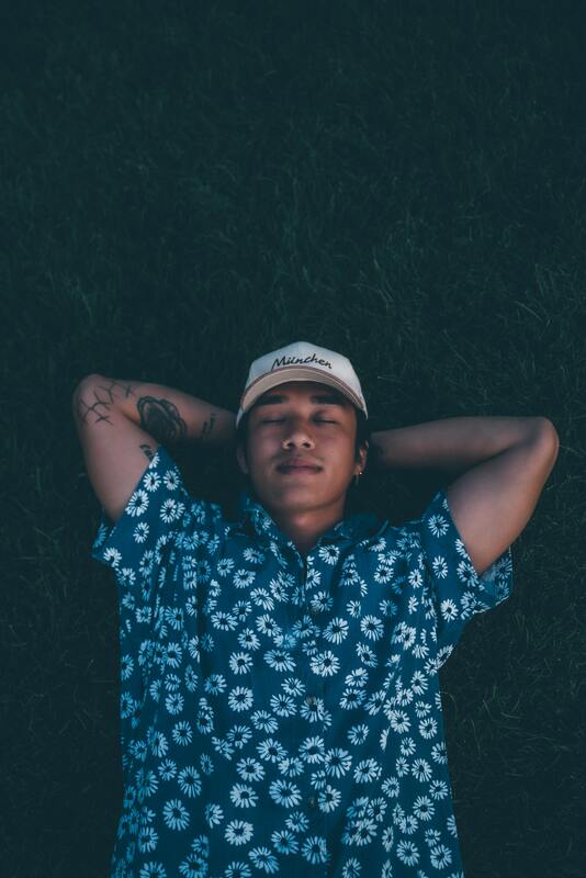 Young man with brown skin and an arm tattoo lies on the ground, eyes closed, and hands folded behind head. This illustrates that Sacred Circle Holistic Healing offers online therapy for young adults who are highly sensitive.