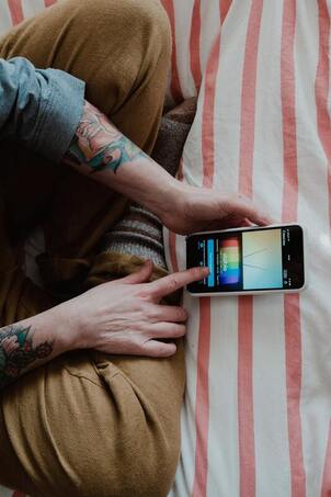 A bird's eye view of a woman's tattooed arms holding an iPhone, legs crossed, sitting on a bed. This illustrates that Sacred Circle Holistic Healing offers counseling online in Kansas for anxiety, Highly Sensitive People, and narcissistic abuse recovery.