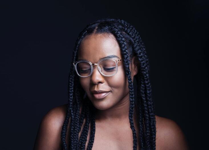 Black woman with clear framed glasses smiling and looking down in front of a black background. This illustrates that Sacred Circle Holistic Healing provides counseling to women who identify as Highly Sensitive People.