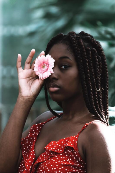 Picture of a black woman holding a pink flower in front of one eye and staring into the camera, not smiling. This illustrates that Sacred Circle Holistic Healing provides therapy in Kansas for women who are struggling with anxiety.