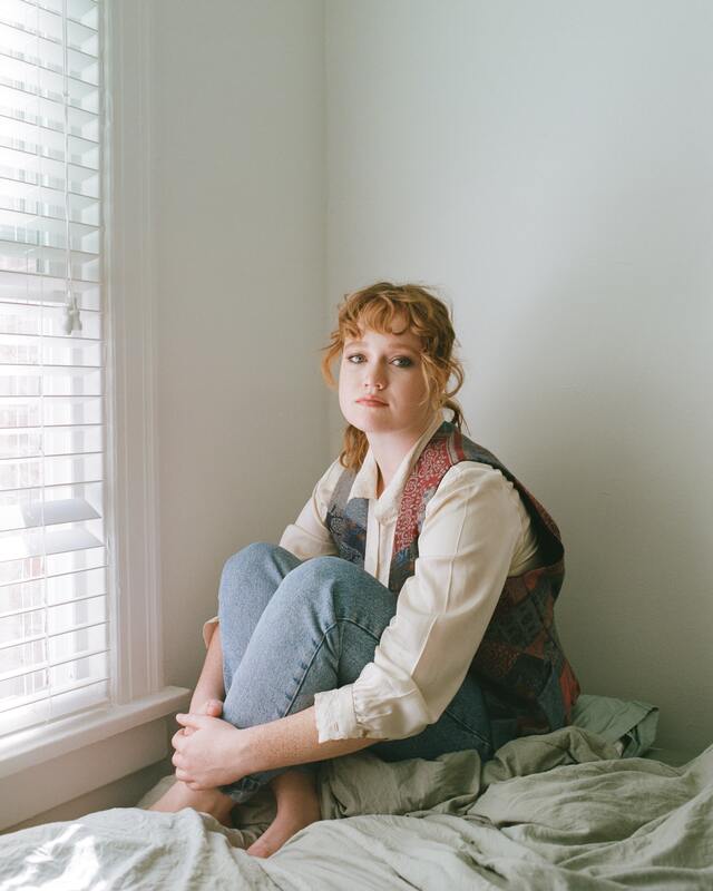Woman with red hair sitting on bed, staring at camera, not smiling. This illustrates that Sacred Circle Holistic Healing offers online counseling and therapy in Kansas for women who struggle with anxiety, low self-esteem, and relational trauma.
