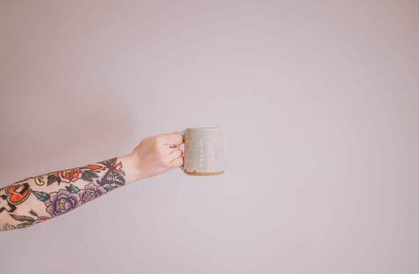 A woman's tattooed arm holding out a mug in front of a white background. This represents being able to sit at home and drink coffee while you do online therapy in Kansas.