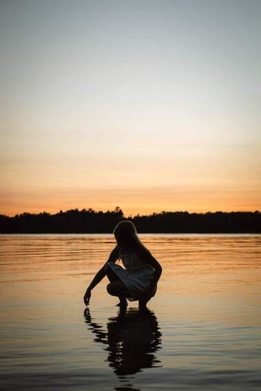 The silhouette of a woman a sunset kneeling down into a body of water. The water illustrates the deep emotions that Highly Sensitive People (HSPs) feel. 
