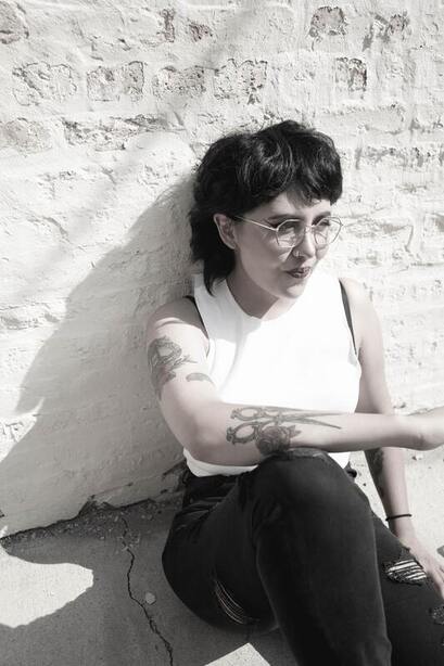 Black and white photo of a woman with short hair, glasses, and a tattoo sitting against a wall. This represents that Sacred Circle Holistic healing offers therapy for women in Kansas recovering for toxic relationships and narcissistic abuse.