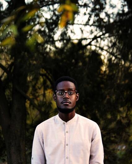 Portrait of a young Black man wearing a button up white shirt and glasses, staring into the camera in front of trees. This illustrates that Sacred Circle Holistic Healing offers specializes in online therapy in Kansas for highly sensitive people.