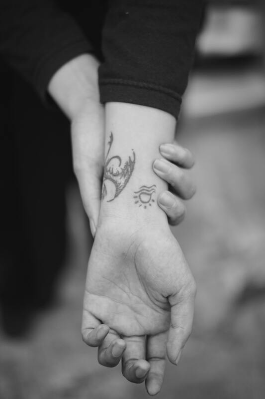 Black and white photo of hands; one is holding the other's wrist with tattoos. This represents the embodied nature of grief. Maggie Jones Boyle offers grief and bereavement counseling in Lawrence, Kansas.