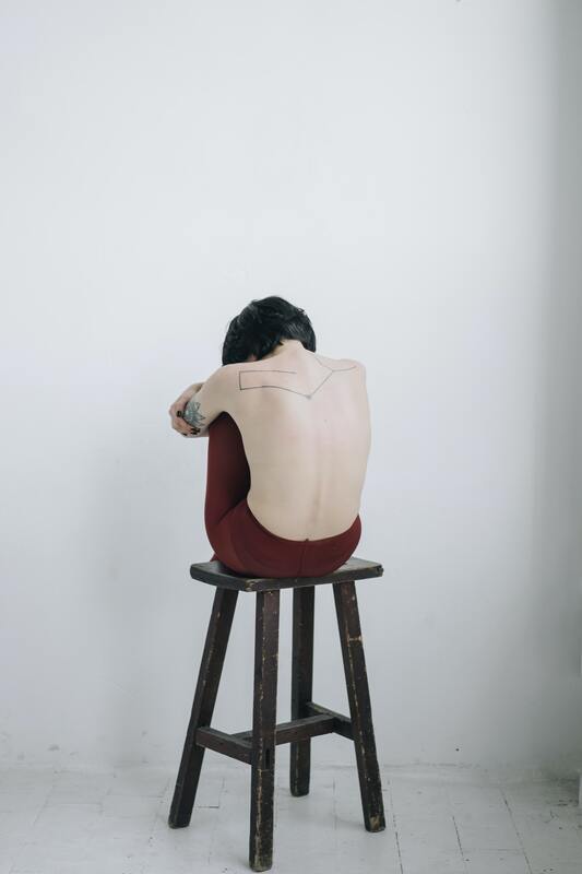 A person with short black hair sitting topless on a stool, minimalist tattoo on their back, back to the camera, in front of a white wall. This illustrates that Sacred Circle Holistic Healing offers online therapy in Kansas for young adults struggling with anxiety, low self-esteem, codependency, and relationship issues.