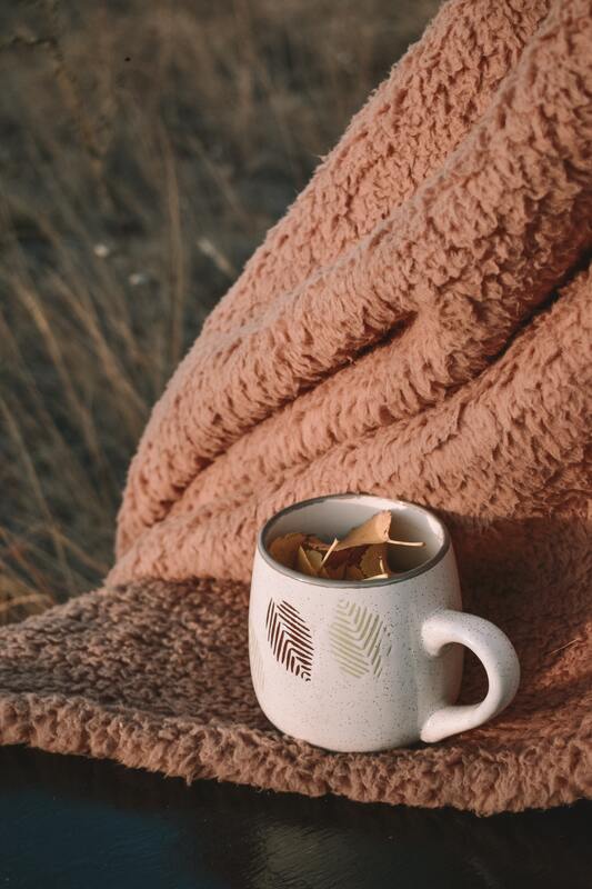 Coffee mug full of yellow leaves, sitting on top of a light brown, fuzzy blanket. This represents that grief rituals can look many different ways. 