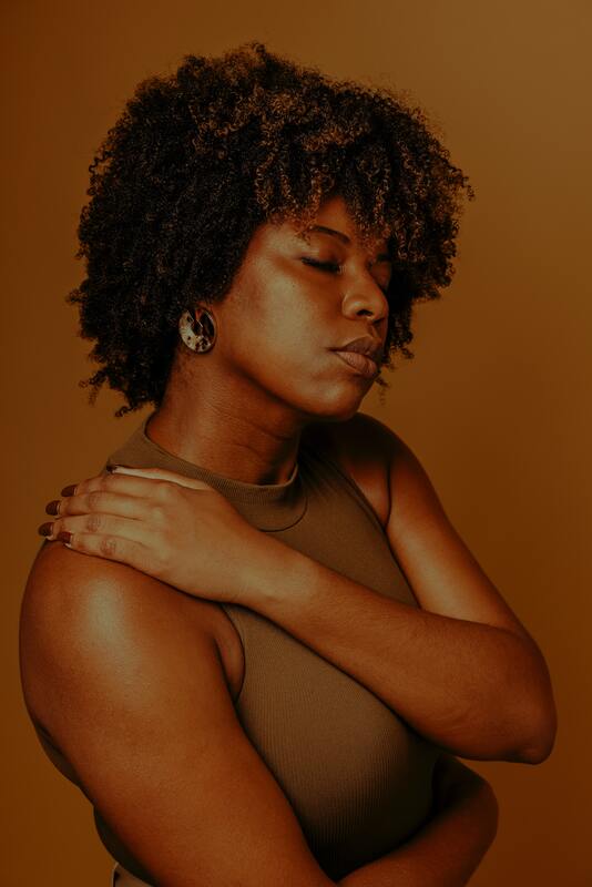 Photo of Black woman in front of a brown wall hugging herself. This represents that Sacred Circle Holistic Healing offers online therapy in Kansas and helps young women love themselves.