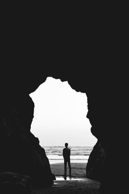 Black and white picture of the silohette of a person standing at the mouth of a large cave. This represents how grief can feel overwhelming. Sacred Circle Holistic healing offers online grief and bereavement counseling in Lawrence, Kansas.